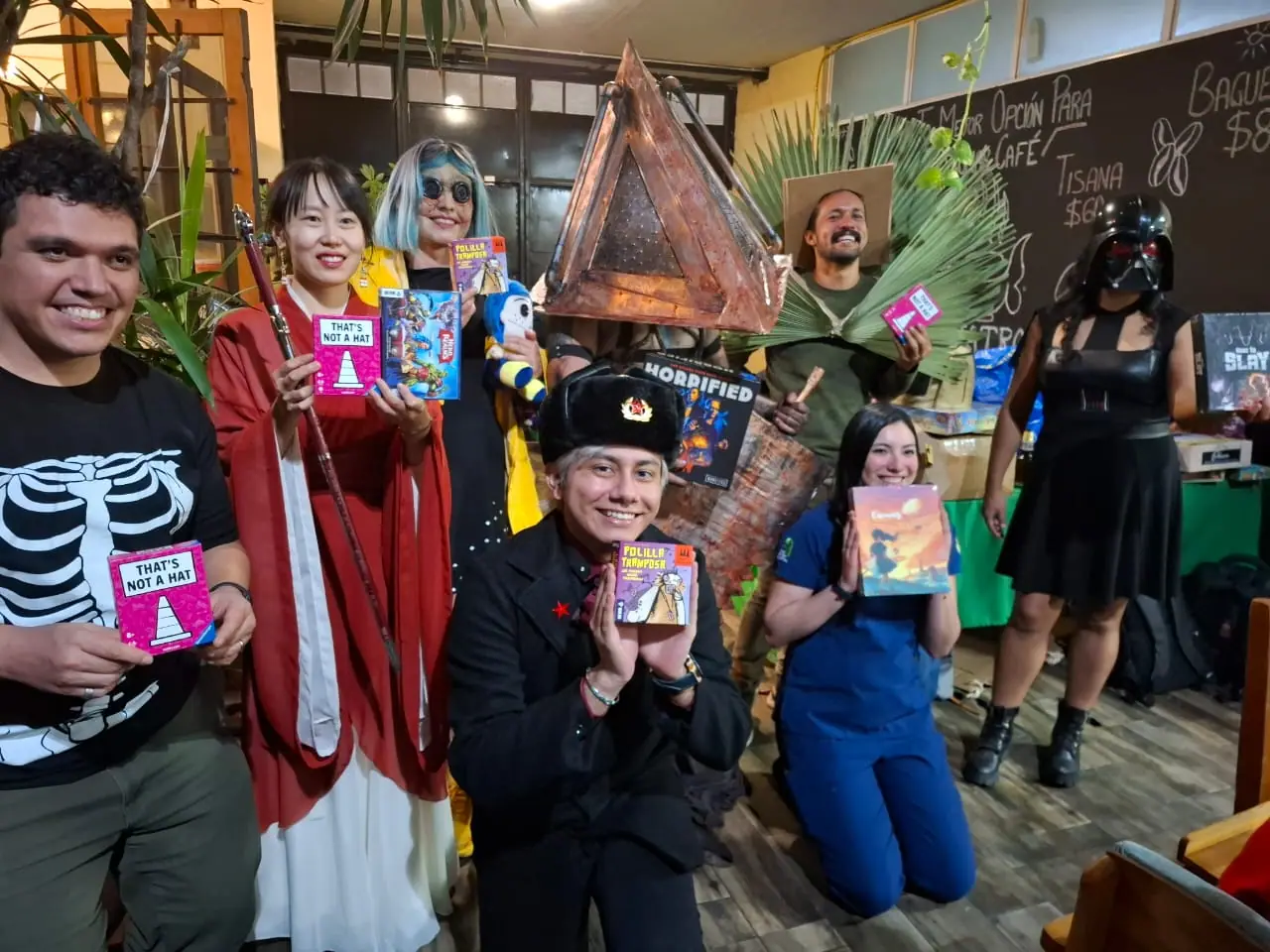 Participantes del concurso de disfraces posan juntos con juegos de mesa en las manos: un gorro ruso, una túnica roja, un casco en forma de pirámide, hojas de palma, un uniforme quirúrgico azul y un casco de Darth Vader.